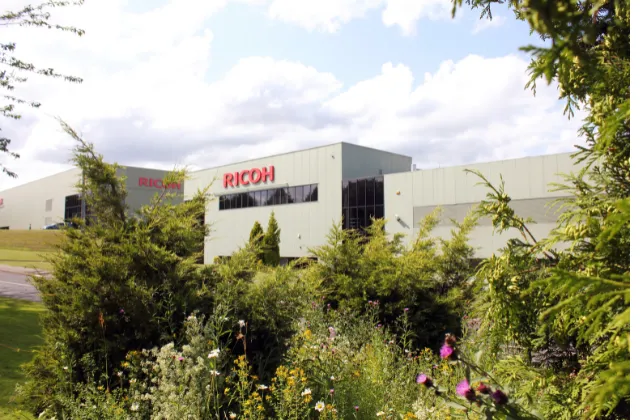 Red fading banner into an outdoor area with greenery in the foreground and the ricoh building in the background