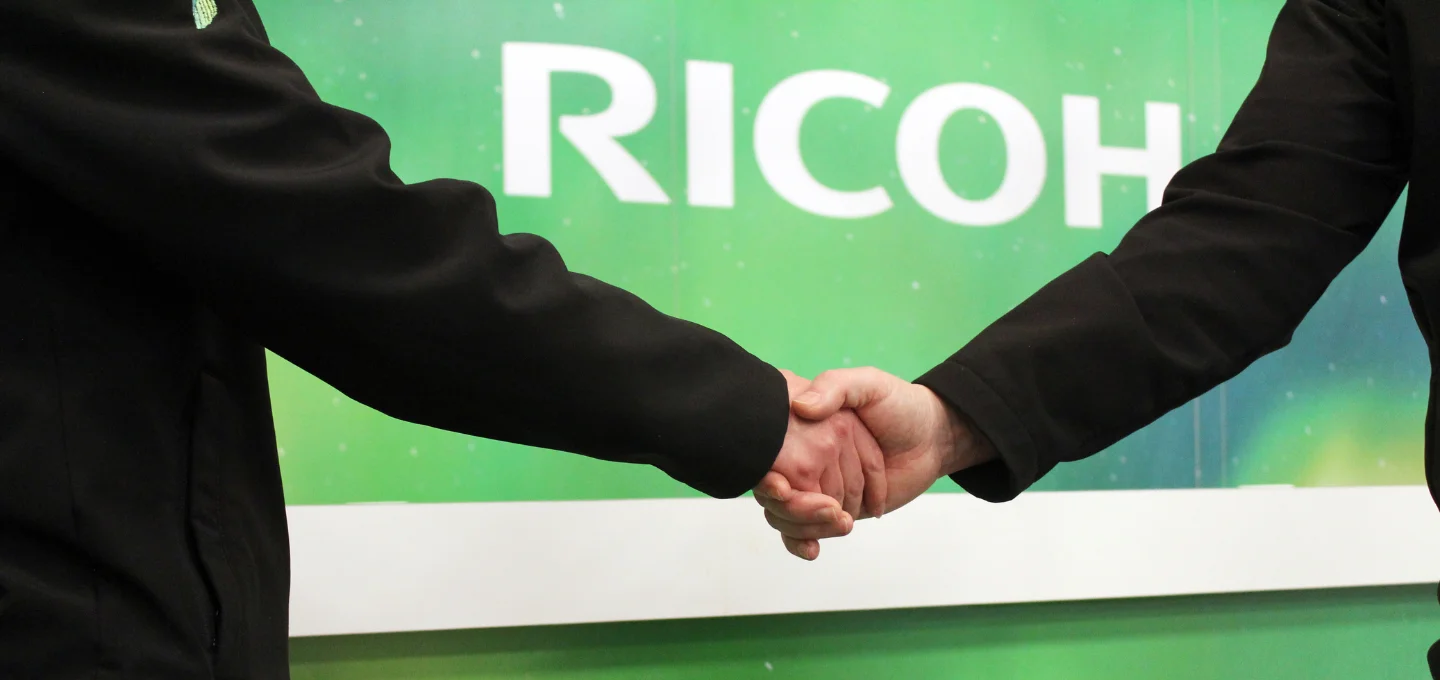 Two individuals shaking hands in front of a Ricoh branded wall and welcome sign.