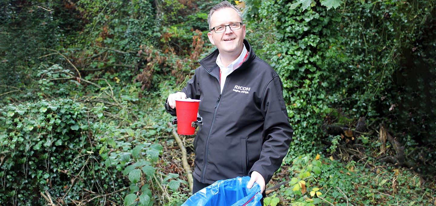 A male employee smiling at the camera, holding a red cup with a litter picker in one hand and a blue rubbish bag in the other