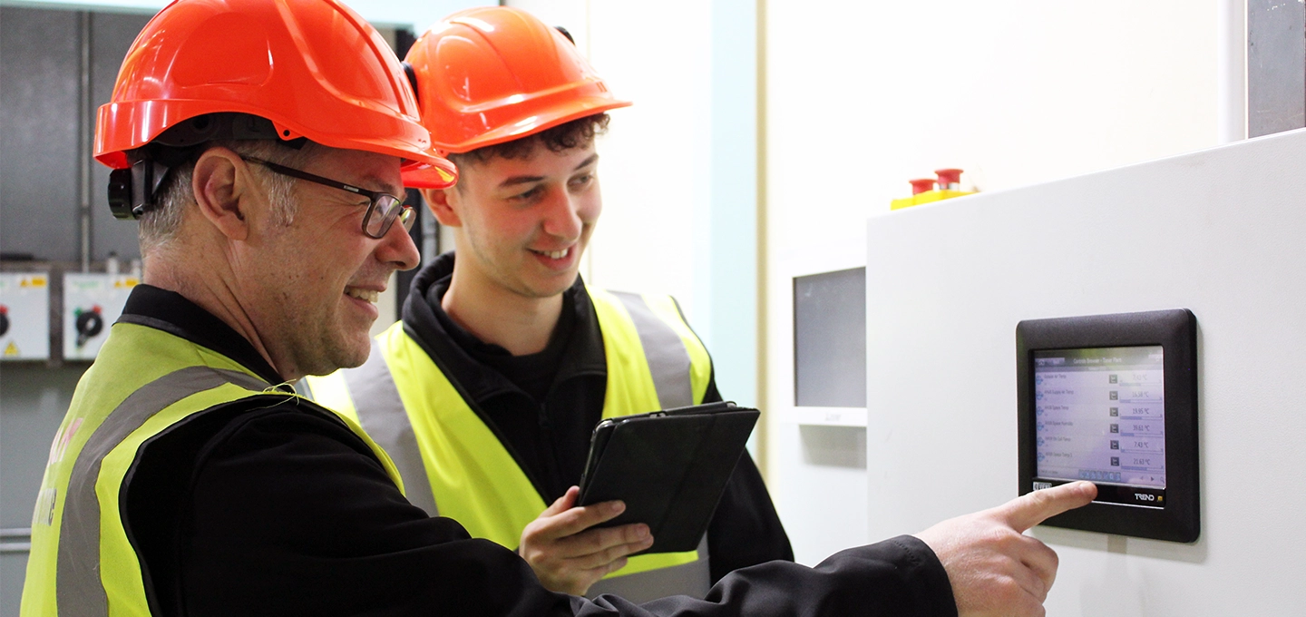 Two employees in PPE including a hardhat and a high vis vest, looking at a control panel